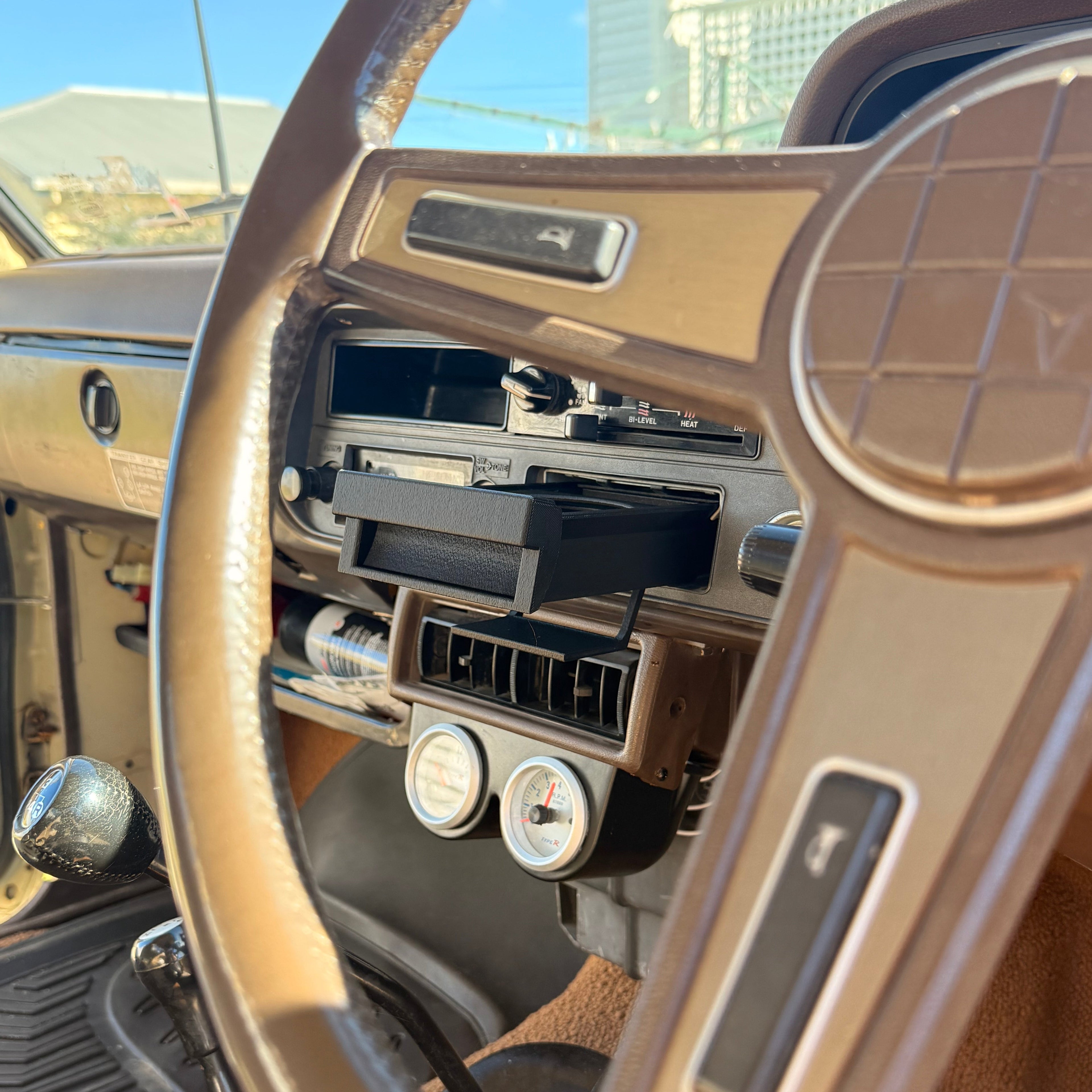 Close-up of a vintage car interior dashboard with a tan steering wheel, brown console, radio, and two round gauges, photographed in bright daylight.