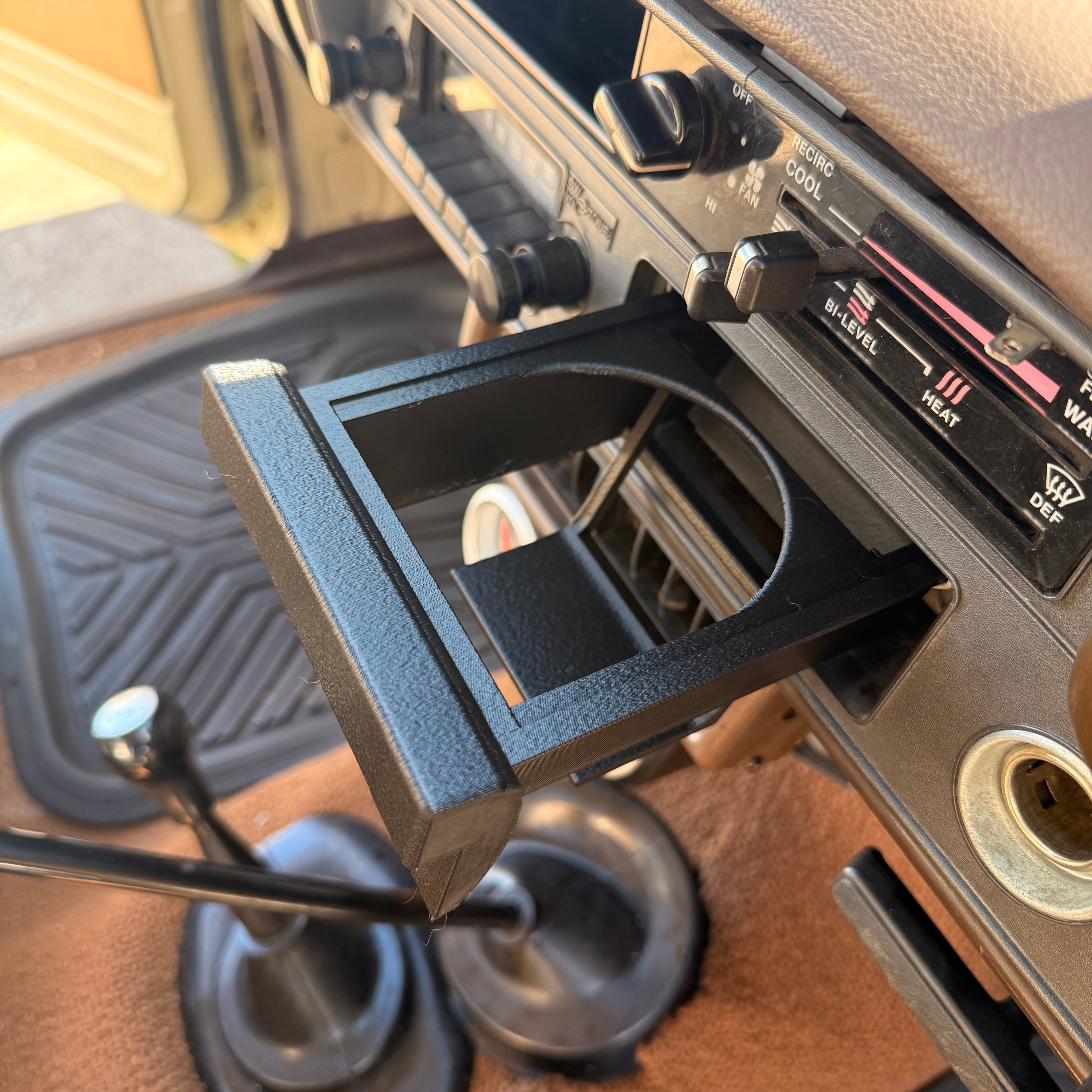 A black cup holder organizer mounted on a car dashboard, shown installed in a vehicle interior with a gear shift stick and footwell visible.