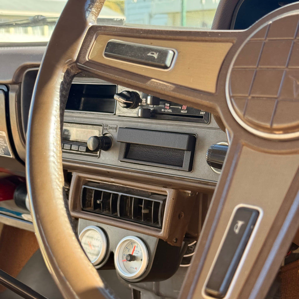 Close-up of a vintage car dashboard and steering wheel, showing tan plastic trim, center console vents, gauges, and a key in the ignition.