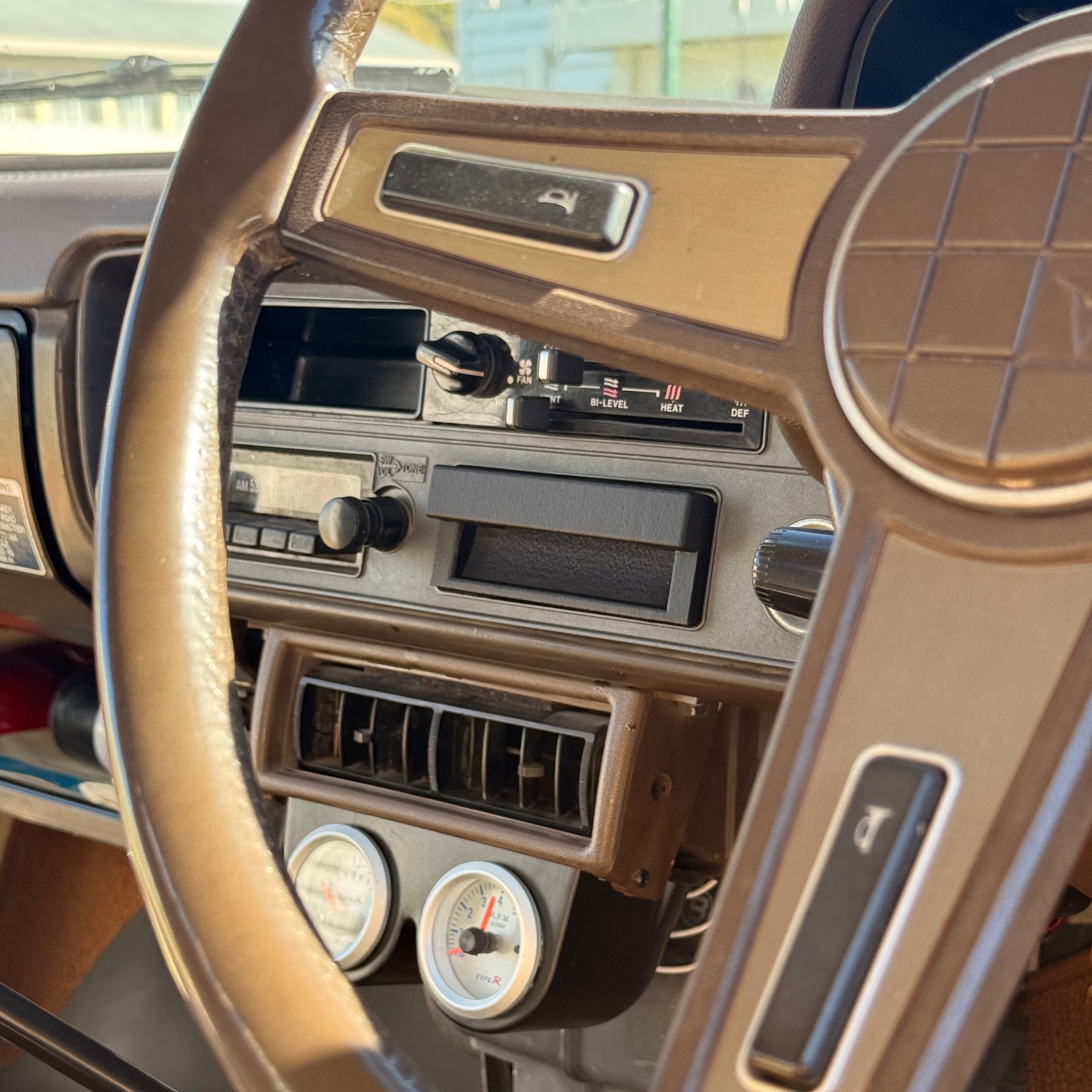 Close-up of a vintage car dashboard and steering wheel, showing tan plastic trim, center console vents, gauges, and a key in the ignition.