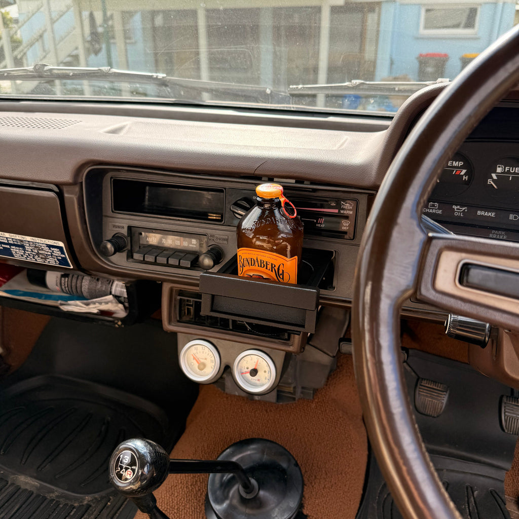 A brown plastic car dashboard accessory holding a bottle of Sam Adams, installed in a vintage vehicle interior with a manual gear shift and steering wheel visible.