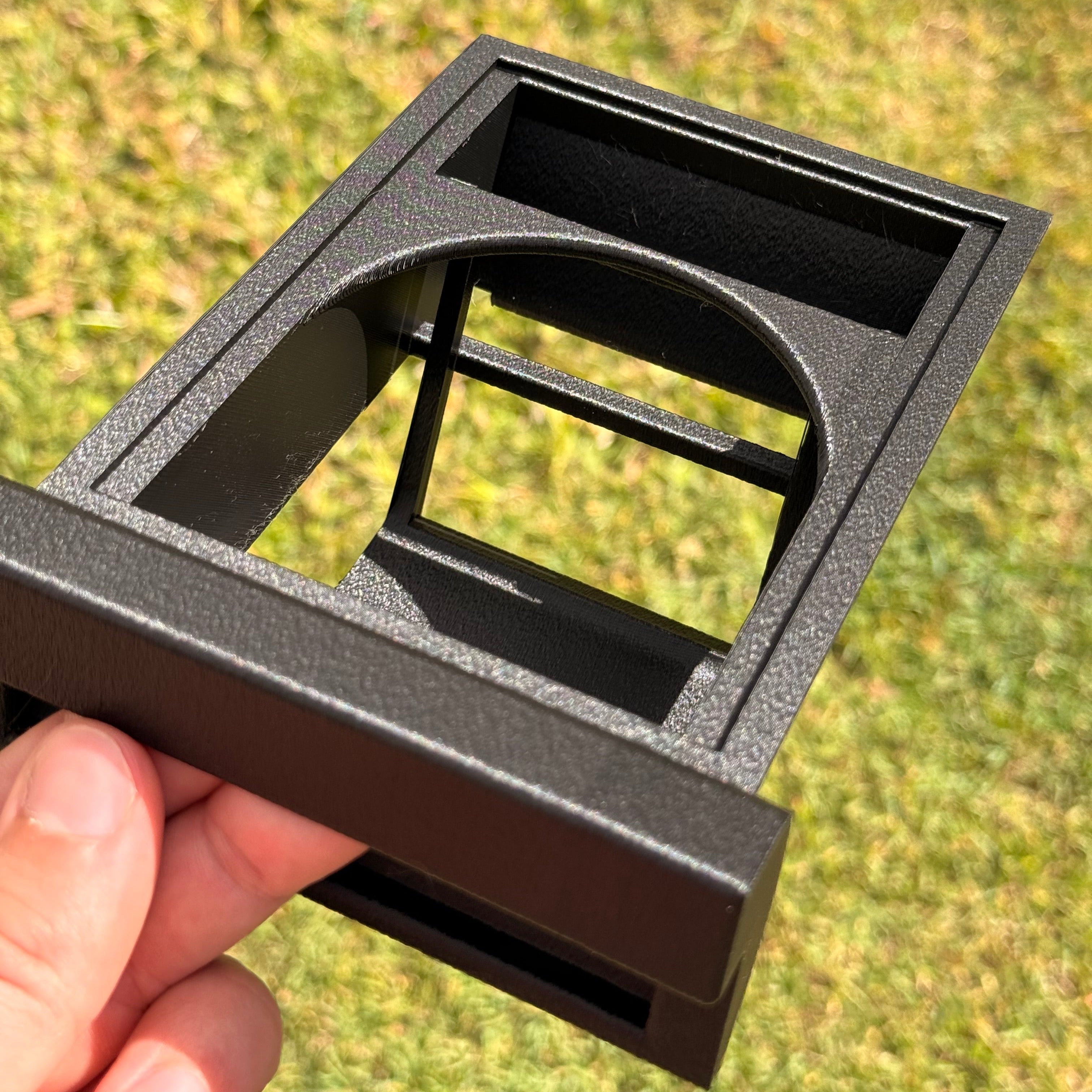 A black textured plastic frame component being held by a hand against a blurred green grass background.